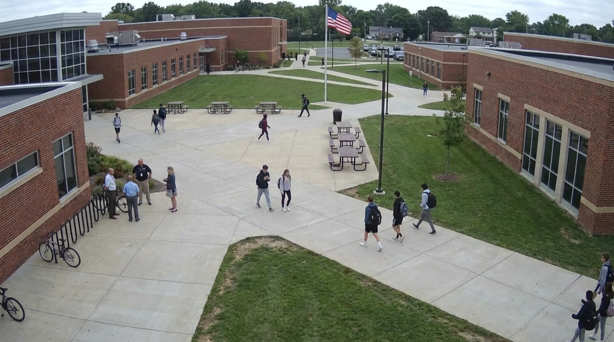 Cloud video surveillance view of a school campus courtyard, showing students and staff moving between buildings with centralized monitoring to support safeguarding and site visibility.