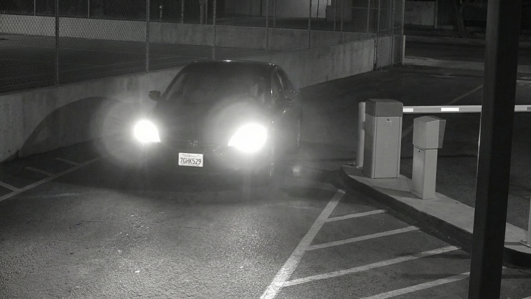 Nighttime security camera view of a car approaching a gated parking entrance, with headlights on and the license plate visible for automatic recognition.